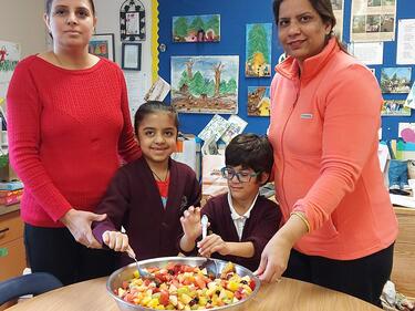 Two adult females helping two student make fruit salad